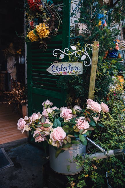 A bouquet of light pink roses with full, lush petals and green leaves rests in a metallic watering can. The arrangement is set outdoors among various green plants and flowers, with a small garden path