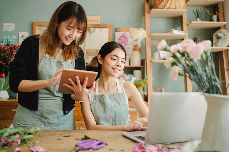 Two women working together in a flower shop, one standing and holding a tablet, and the other sitting at a wooden table surrounded by floral materials. The seated woman, with dark hair, is smiling at 