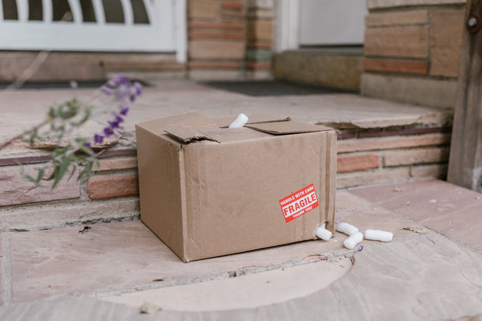 A cardboard delivery box with a red 'Fragile' label is placed on outdoor brick steps near a door. The box appears slightly damaged with some creases on the top. A few white packing peanuts are scatter