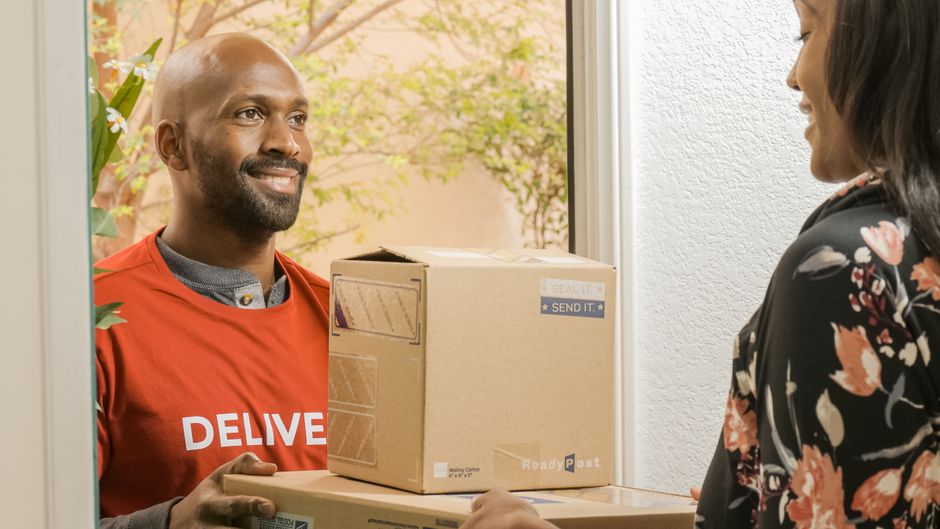 A floral delivery courier from Belvedere Flowers, wearing a red delivery vest with 'DELIVE' visible, hands over a cardboard package to a woman at her door. The woman, dressed in a black floral blouse,