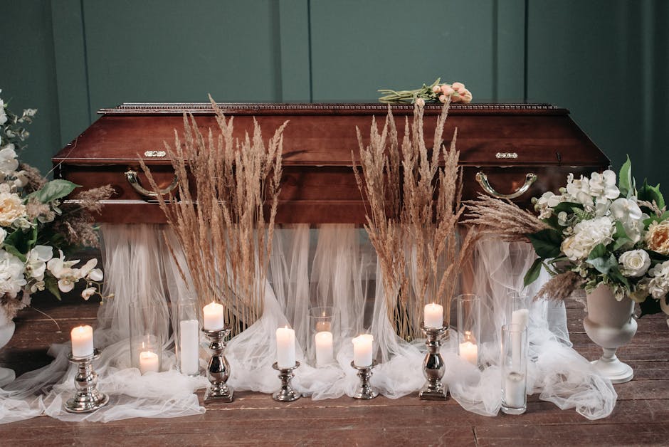 A wooden casket is adorned with white and cream floral arrangements, including roses and other delicate flowers, placed in white vases on either side. Tall, dried pampas grass stems are arranged uprig