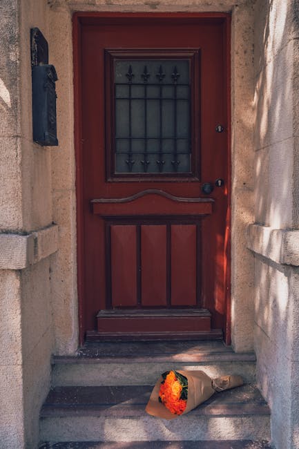 A bouquet of fresh flowers resting on stone steps in front of a deep red wooden door with a black iron grille window. The bouquet includes orange and yellow flowers, wrapped in brown paper, with visib