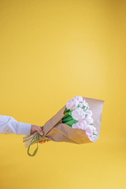 A person’s hand is visible holding a bouquet of pastel pink tulips, wrapped in light brown paper with a green ribbon, against a vibrant yellow background. The tulips have fresh, smooth petals and gr
