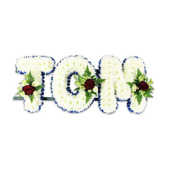 A carefully handcrafted funeral tribute that spells TOM in bold, three-dimensional letters created from a multitude of small white chrysanthemum blooms. The chrysanthemums are densely placed to form a soft, pillowy texture that invites close inspection; their clean white tones convey purity and quiet comfort. Each letter is edged with a vivid royal blue ribbon, its saturated colour providing a crisp, formal outline and a striking contrast to the white surface. Incorporated into the centre and lower areas of each letter are composed floral sprays: a solitary, deep red rose serves as the focal point within each cluster, symbolising enduring love and respect, while surrounding ivory spray roses and tight white buds lend delicate nuance and a sense of care. Slender fern fronds and rich green foliage add vertical movement and natural texture, balancing the sympathy palette with refreshing green highlights. A subtle dark metal support runs along the base, visible beneath the letters, revealing the practical armature that keeps the tribute upright during a service. Photographed against a bright white background with soft, even illumination, the arrangement reads clearly and respectfully, the blue ribbon's sheen and the rose's velvety richness contrasting beautifully with the chrysanthemum's matte softness. This personalised piece is fitting for memorial services in Belvedere and neighbouring communities-perhaps placed near a riverside chapel or on a church stand-offering a composed, comforting expression of honour and remembrance.