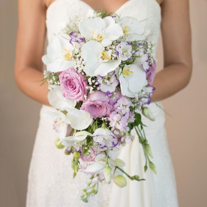 In the heart of Belvedere, a bride stands in elegant repose, her white strapless gown adorned with subtle lace details as she presents an extraordinary cascading bouquet against a warm, softly faded background. The floral design showcases luxurious white phalaenopsis orchids, their stately forms and golden centers complemented by the pillowy texture of blush-pink garden roses and feathered accents of lavender and light purple lisianthus. Stock flowers, subtle and spiky, nestle between the larger blooms, adding movement and gentle contrast alongside airy white gypsophila and hints of green foliage. The composition's graceful teardrop fall captures the dynamic yet serene spirit of a Belvedere celebration-easily imagined in venues near local parks or riverside chapels. The bouquet's delicate fresh fragrance pairs with an understated elegance, inviting onlookers into a scene of romance and hope. This image beautifully demonstrates Belvedere Flowers' mastery of timeless wedding styles, grounded in local tradition and modern artistry.