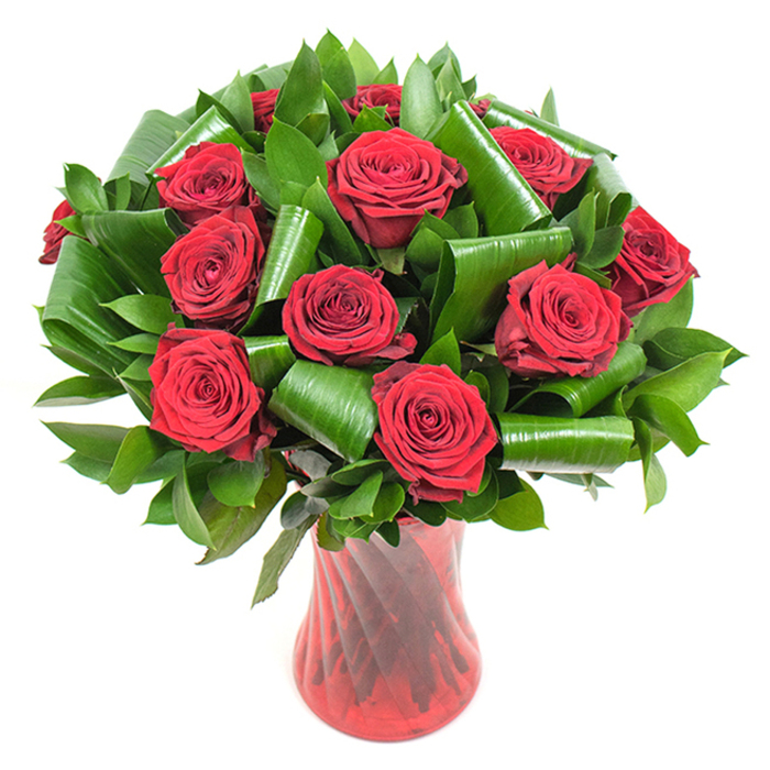A luxurious, hand-composed bouquet of roughly twelve to fifteen deep crimson roses sits poised in a tall, ruby-red glass vase, the kind of arrangement that reads as both lush and deliberately refined. Each rose displays perfectly formed, velvety petals that spiral outward from a tight core, their surfaces catching gentle, diffused light to reveal subtle tonal shifts within the red - hints of burgundy and ruby that lend the blooms tactile depth. Supporting the roses is abundant dark-green foliage: narrow glossy leaves forming a dense backdrop and broader, emerald leaves that have been artfully rolled and curled into tubular shapes and sweeping loops. These sculpted green elements create an architectural frame that contrasts the softness of the petals and adds modern structure to a timeless flower. The vase is tall and gracefully tapered, its translucent red glass showing a faint ribbed or twisted texture which throws vertical highlights and reflections that mirror the bouquet's passionate palette. Placed against a clean white background, the arrangement becomes an isolated study in colour, texture and form - perfect for an anniversary table, a Valentine's display, or an elegant gift meant to convey deep affection. There is a sensory suggestion of a classic rose scent, subtle and lingering, and the whole piece feels like something a skilled local florist might prepare for same-day delivery across Belvedere, Erith or the nearby riverside neighbourhoods - a considered expression of love and refined taste executed with steady craftsmanship.
