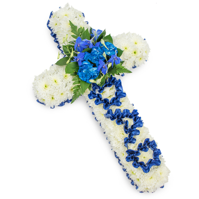 In this image, a full floral cross is laid out on a bright white surface, the high contrast drawing attention to its colour and craft. The cross shape is completely filled with small white spray chrysanthemums, each bloom turned outward so that only layers of overlapping petals are visible. This mass of flowers creates a plush, consistent texture, reminiscent of soft fabric or snow, and conveys a sense of quiet purity. Tracing all around the outer edge, a band of royal blue satin ribbon adds structure, its smooth, slightly glossy finish catching the light and defining the cross with a stately border. At the core where the arms of the cross intersect, a lush central spray of blue flowers has been arranged. Bold blue carnations or similarly dyed blooms are clustered at the centre, while finer, elongated florets reminiscent of blue delphiniums or orchids rise gently from within the group, providing height and intricate detail. Interspersed with these blue tones are rich green fern fronds and other foliage, whose pointed and rounded leaves introduce an organic feel, as though a small bouquet has been set into the cross. Running down the main vertical bar, the word DAD appears, not printed but crafted from the same royal blue ribbon, folded and looped into three clear letters that stand out against the white floral base. This handmade lettering signals that the piece is intended as a tribute for a father, adding emotional weight to the design. Lit evenly from above, with just a soft shadow around the edges, the cross looks substantial and carefully constructed. It is the kind of thoughtful memorial arrangement a family in Belvedere might choose to have delivered from Belvedere Flowers to accompany a church service or cremation, offering a visible sign of respect and lasting love.