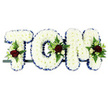 Here, the camera focuses on a bespoke floral tribute that spells out the name TOM in prominent block capital letters, often chosen for funerals and memorial services in Belvedere. The letters are arranged horizontally on a slender support bar and photographed against a simple white background, which allows every texture and colour to stand out clearly. Each letter is densely covered with pristine white chrysanthemums, their small blooms clustered tightly until they form a soft, pillowed surface that looks comforting to the touch. Tracing the outer edge of each capital is a royal blue satin ribbon, its smooth sheen catching the light and providing a crisp outline that enhances the legibility of the name. At thoughtful intervals - near the upper left of the T, along the right side of the O, and at the top right of the M - there are tasteful clusters of accent flowers. These feature deep crimson or burgundy roses, creamy white spray roses, and elegant white flowers reminiscent of freesia, all woven among fresh green foliage and delicate fern fronds. The bold contrast of red, white, blue, and green suggests emotions of love, purity, and gentle remembrance. Although the setting is minimal, one can imagine this tribute carefully delivered by Belvedere florists to a local chapel or cemetery, the faint fragrance of chrysanthemums and roses mingling with the stillness of the service. It is a personalised arrangement that honours Tom by name, providing a clear, heartfelt focal point for family and friends to reflect on as they say their final goodbyes.