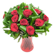 A rich, rounded bouquet of twelve or more deep crimson roses fills the frame, each bloom at the peak of its velvety, spiralled perfection. The petals are a wine-dark red, their surfaces catching soft, even studio light so you can sense the plush texture and delicate edges of every tightly layered curl. Dark green foliage forms a generous collar around the roses - flat, glossy leaves that read like Salal or Ruscus create a dense, verdant backdrop while broader, emerald-gleaming leaves are artfully folded and rolled into elegant cylinders and dramatic loops. Those sculpted leaves arc and spiral like small architectural gestures, lending the classic roses a modern, structured silhouette and breathing depth into the composition. The stems sit in a tall, translucent ruby-red glass vase whose slender profile, gently flared rim and subtly twisted ribbed surface throw vertical highlights that echo the roses' passionate tones. Against a crisp white background the intense reds and lush greens pop in high contrast, isolating every curve, fold and shadow so the arrangement reads as a single, composed statement. There is an implied scent of soft classic rose perfume - warm, slightly honeyed and quietly persistent - that seems to lift from the clustered heads. This is a bouquet that balances traditional romance with contemporary form; perfect as an anniversary or Valentine's centrepiece, a heartfelt gift, or a dramatic dining table focal point. For Belvedere customers it reads like a familiar, handcrafted signature piece you might spot at the town's market or a window on the High Street, a floral language of love rendered with confident technique and calm restraint.