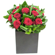 A modern, sculptural bouquet of deep crimson roses sits in a matte black square box, a composition that reads equal parts romance and urban calm. Each rose opens in velvety spirals, the petals catching a steady, clear light so the crimson appears almost luminous against the dark vessel. Broad, glossy green leaves have been carefully curled and layered around the blooms, their waxy sheen and rhythmic loops creating movement and depth so the cluster reads like a living relief. The square cube grounds the design with a confident, minimal silhouette, its matte finish absorbing light and setting off the roses' plush texture. From close by you would sense a light, clean perfume-the classic sweetness of rose softened by fresh, almost green notes from the foliage, a subtle contrast between heady romance and crisp freshness. This arrangement was composed with intention; it suits an anniversary table, a heartfelt declaration, or a sleek office reception in and around Belvedere and nearby Erith. The craftsmanship shows in the way each leaf cradles a bloom and in the balanced negative space that lets the bouquet breathe. As a florist who often works for customers collecting from Belvedere station or arranging hand-deliveries to local addresses, I picture this design brightening a riverside flat by the Thames or a contemporary dining table near Crossness. It's the kind of piece that reads beautifully in photos and even more so in person-the visual contrast of matte and velvet, the tactile invitation of soft petals and glossy foliage-a quietly bold statement that lingers both visually and in scent.
